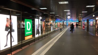 Corredor movimentado de um shopping center, com consumidores, lojas e um relógio em destaque.