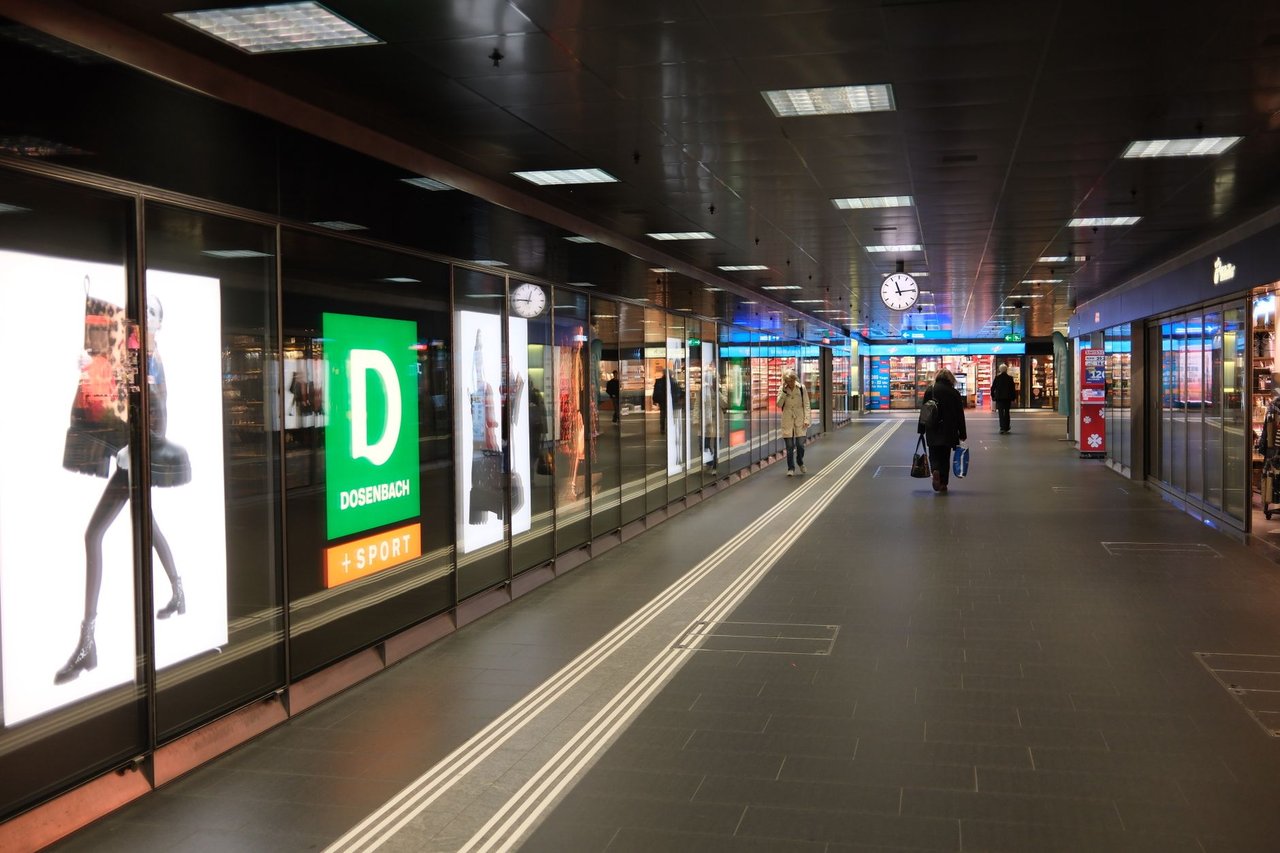 Corredor movimentado de um shopping center, com consumidores, lojas e um relógio em destaque.