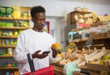 Homem examina uma manga enquanto faz compras e consulta o celular em um supermercado.