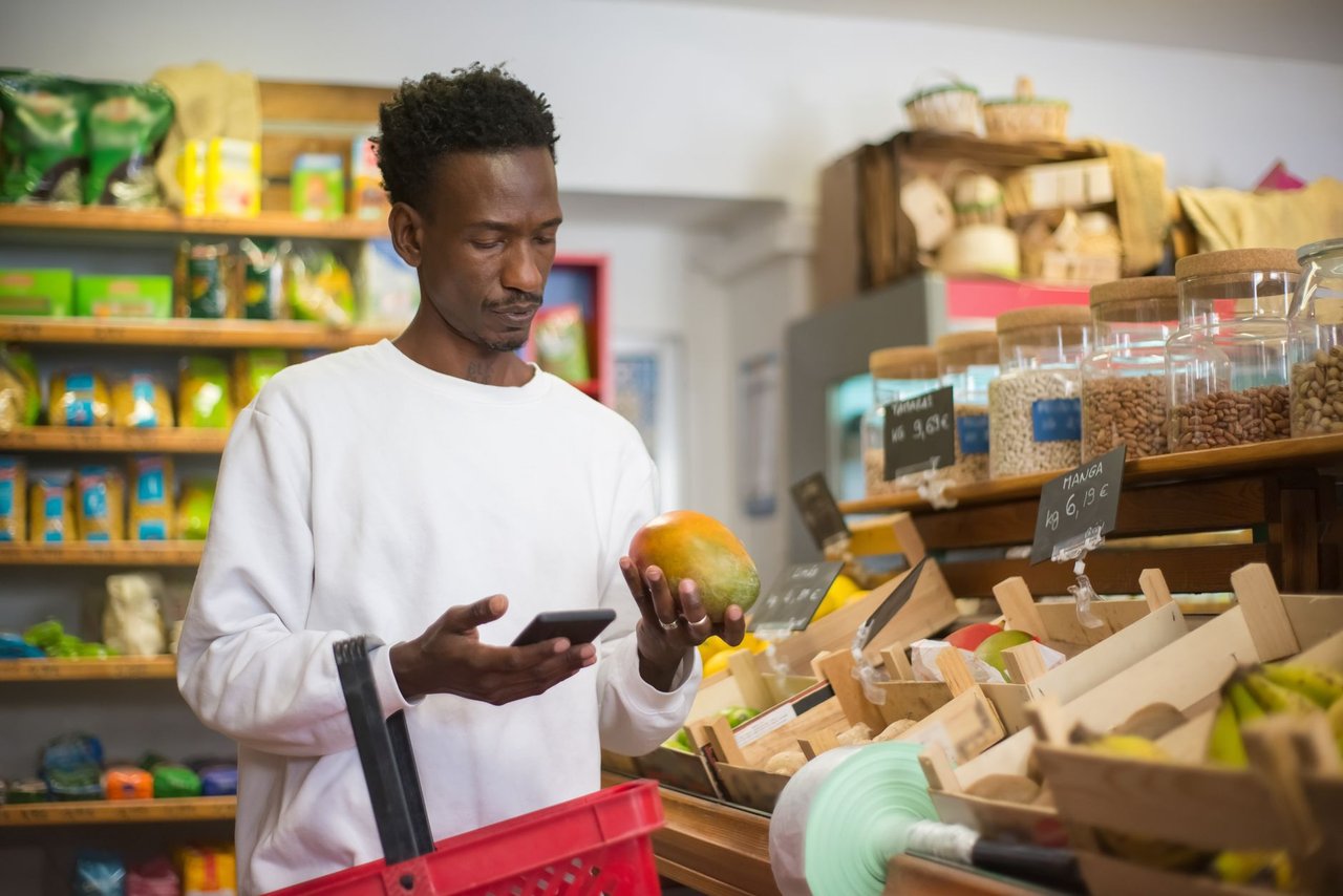 Homem examina uma manga enquanto faz compras e consulta o celular em um supermercado.